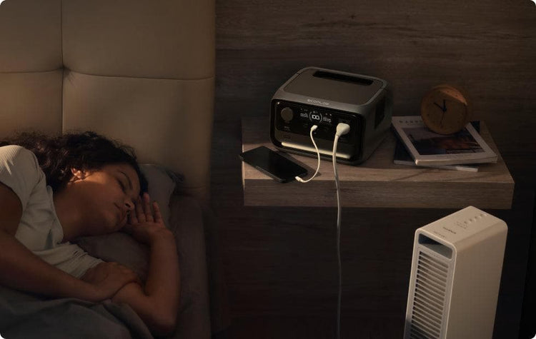 Mulher dormindo ao lado de uma estação de energia portátil EcoFlow, que está carregando um smartphone e alimentando um aquecedor elétrico no quarto, destacando seu funcionamento silencioso e seguro.